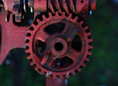 A close-up view of a rusty red gear with intricate teeth and a central hole. The gear is part of a larger mechanical assembly with additional metallic components visible in the background. The overall texture is rough with visible signs of wear and rust.