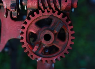 A close-up view of a rusty red gear with intricate teeth and a central hole. The gear is part of a larger mechanical assembly with additional metallic components visible in the background. The overall texture is rough with visible signs of wear and rust.
