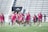A group of women wearing bright pink athletic uniforms with matching shoes walks across a sports field. Each carries a lacrosse stick and has a sports bag. They appear focused and are moving together in a coordinated manner. Stadium seating and a sign labeled 'MEN'S RESTROOM' are visible in the background.
