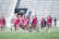 A group of women wearing bright pink athletic uniforms with matching shoes walks across a sports field. Each carries a lacrosse stick and has a sports bag. They appear focused and are moving together in a coordinated manner. Stadium seating and a sign labeled 'MEN'S RESTROOM' are visible in the background.