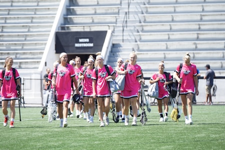 A group of women wearing bright pink athletic uniforms with matching shoes walks across a sports field. Each carries a lacrosse stick and has a sports bag. They appear focused and are moving together in a coordinated manner. Stadium seating and a sign labeled 'MEN'S RESTROOM' are visible in the background.
