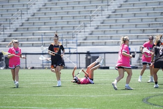 A group of female lacrosse players are actively engaged in a game on a grassy field. One player is seen in mid-air, having tripped or fallen, while the others are either running or standing nearby. Some players are wearing pink jerseys, and others are in darker uniforms. Stands with empty seats are visible in the background, indicating a sports stadium setting.