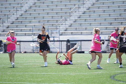 A group of female lacrosse players are actively engaged in a game on a grassy field. One player is seen in mid-air, having tripped or fallen, while the others are either running or standing nearby. Some players are wearing pink jerseys, and others are in darker uniforms. Stands with empty seats are visible in the background, indicating a sports stadium setting.