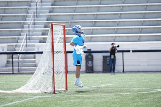 A lacrosse player in a bright blue uniform and helmet is leaping in the air next to a lacrosse goal on a grassy field. The background includes empty bleachers and a person with a camera positioned to capture the action.