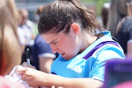 A person with dark hair is wearing a light blue Adidas top and appears focused on signing an object with a black marker. The setting is outdoors with other people visible in the background.