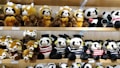 Multiple rows of plush toy pandas and red pandas are arranged neatly on wooden shelves. The toys wear various accessories, like graduation caps and striped shirts. The scene features a mix of brown and white colors, along with black and white on the panda toys.