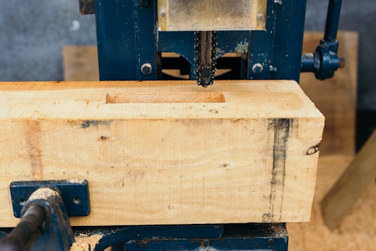 A woodworking machine is partially visible, with a large wooden beam being processed. The machine's metal components are visible, with sawdust scattered around. The setting appears to be a workshop with other wooden pieces in the background.