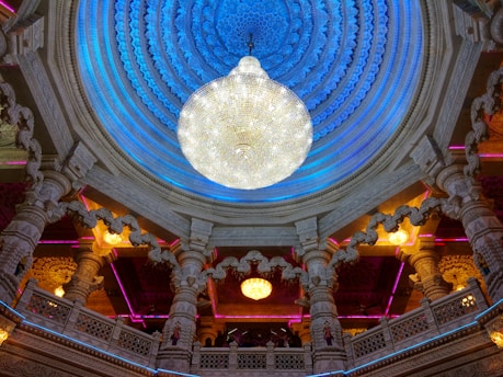 An intricately designed interior of a structure featuring a large, ornate chandelier hanging from a domed ceiling. The ceiling is decorated with elaborate blue-toned patterns and surrounded by intricate architectural details, including carved columns and arches with warm lighting accents.