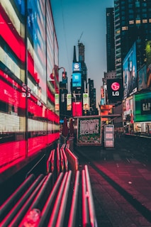 Bright neon lights and digital billboards dominate a bustling urban area. The reflections of vibrant red and blue lights create an energetic atmosphere. Large advertisements and signs are prominently displayed on the buildings, including well-known brands and a prominent sign inviting people to 'Discover Broadway's Secrets'. The setting is a city center, likely a famous location.