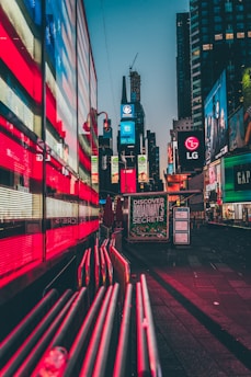 Bright neon lights and digital billboards dominate a bustling urban area. The reflections of vibrant red and blue lights create an energetic atmosphere. Large advertisements and signs are prominently displayed on the buildings, including well-known brands and a prominent sign inviting people to 'Discover Broadway's Secrets'. The setting is a city center, likely a famous location.