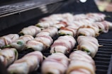 Several bacon-wrapped items are neatly placed on a grill, each secured with a toothpick. The bacon appears to be uncooked or partially cooked, with a pink and white marbled texture. The green inside suggests the item might be jalapeño poppers or similar stuffed vegetables.
