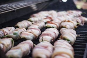 Several bacon-wrapped items are neatly placed on a grill, each secured with a toothpick. The bacon appears to be uncooked or partially cooked, with a pink and white marbled texture. The green inside suggests the item might be jalapeño poppers or similar stuffed vegetables.