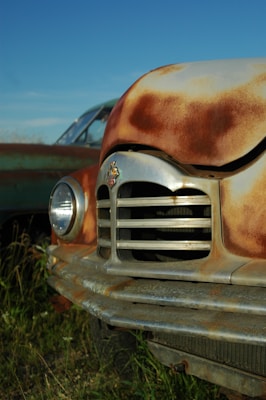 A close-up view of the front of an old, rusty car with a chrome grille and round headlight. The car's body shows signs of age and weathering with patches of rust. In the background, another vintage car is partially visible surrounded by grass under a clear blue sky.