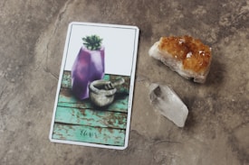 A tarot card is placed on a stone surface, depicting a purple crystal vase with a small green succulent on a teal wooden background. Nearby, there is an orange-colored crystal cluster and a clear quartz point.