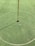 A golf hole on a putting green with a white golf ball near the hole. A red flagstick is positioned in the hole, casting a shadow on the neatly manicured grass. A circular marking surrounds the hole.