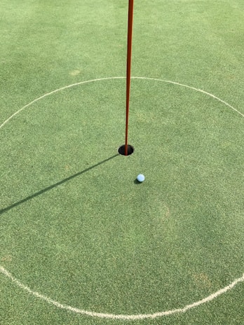 A golf hole on a putting green with a white golf ball near the hole. A red flagstick is positioned in the hole, casting a shadow on the neatly manicured grass. A circular marking surrounds the hole.