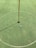 A golf hole on a putting green with a white golf ball near the hole. A red flagstick is positioned in the hole, casting a shadow on the neatly manicured grass. A circular marking surrounds the hole.
