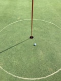 A golf hole on a putting green with a white golf ball near the hole. A red flagstick is positioned in the hole, casting a shadow on the neatly manicured grass. A circular marking surrounds the hole.