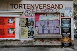 A wall covered with a variety of posters, graffiti, and street art. There are posters for events like 'West Side Story' and 'Deep Web' alongside graffiti art with bold letters and abstract designs. The words 'Tortenversand' are prominently displayed above the art, and there are some recognizable brand logos and text in different languages.