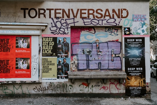 A wall covered with a variety of posters, graffiti, and street art. There are posters for events like 'West Side Story' and 'Deep Web' alongside graffiti art with bold letters and abstract designs. The words 'Tortenversand' are prominently displayed above the art, and there are some recognizable brand logos and text in different languages.