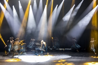 Blurred dancers are captured in motion on a dimly lit stage with bright spotlights shining down, creating a dynamic and ethereal atmosphere. The figures appear to be moving fluidly, with trails of light accentuating their movement.