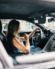 A woman with long, light brown hair is seated in the driver's seat of a luxury car. She is turning the steering wheel, suggesting she might be driving or about to drive. The interior of the car features sleek, modern designs with a focus on dark colors and advanced technology. Through the window, part of a building and outdoor environment is visible, indicating an urban setting.