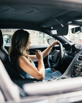 A woman with long, light brown hair is seated in the driver's seat of a luxury car. She is turning the steering wheel, suggesting she might be driving or about to drive. The interior of the car features sleek, modern designs with a focus on dark colors and advanced technology. Through the window, part of a building and outdoor environment is visible, indicating an urban setting.