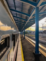 A railway station platform with a train parked alongside. The platform is partially covered by a blue metal and glass canopy, with support beams running along its length. The sky in the background shows a sunset with prominent clouds, casting a warm light on the platform.