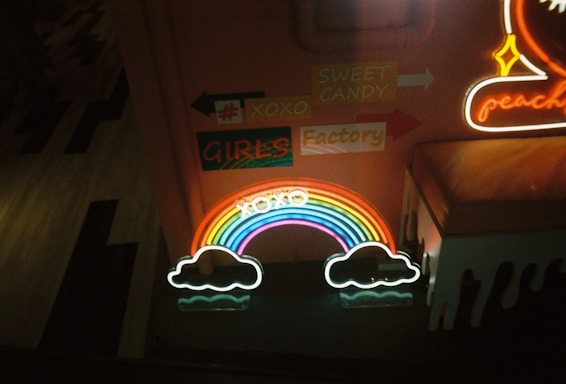 Neon signs displaying various elements such as a rainbow with clouds, directional signage with words like 'SWEET CANDY' and 'GIRLS Factory', and other decorative elements in a dimly lit indoor setting.