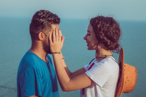 A woman playfully covers the eyes of a man standing beside a calm sea. They are outdoors, with the sea visible in the background. The woman has curly hair and is smiling softly while interacting with the man. She wears a white shirt and holds a straw hat. The man is wearing a blue shirt.