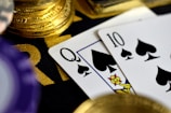 A close-up view of playing cards, specifically a Queen and a Ten of spades, surrounded by shiny gold coins and part of a poker chip, suggesting a gambling or casino theme.