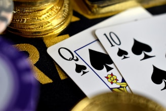 A close-up view of playing cards, specifically a Queen and a Ten of spades, surrounded by shiny gold coins and part of a poker chip, suggesting a gambling or casino theme.