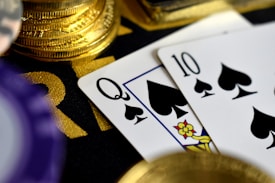A close-up view of playing cards, specifically a Queen and a Ten of spades, surrounded by shiny gold coins and part of a poker chip, suggesting a gambling or casino theme.