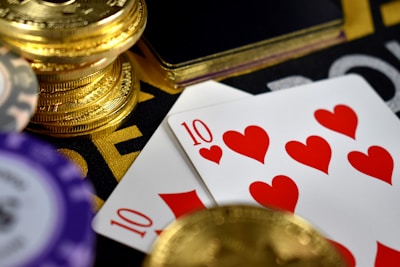A close-up view of playing cards showing the ten of hearts placed alongside poker chips and a stack of gold coins. The scene suggests a gambling or casino setting with high stakes involved.