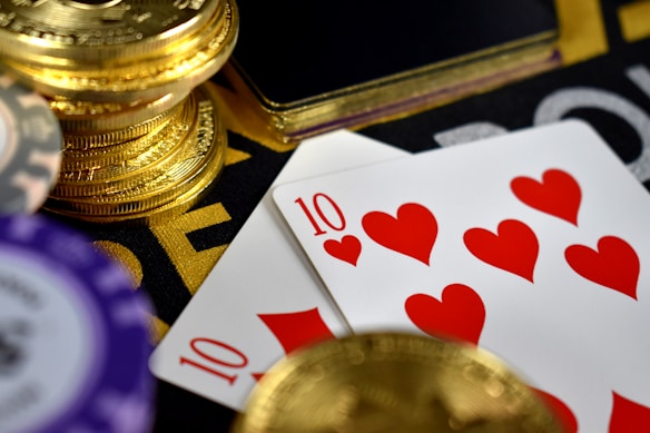 A close-up view of playing cards showing the ten of hearts placed alongside poker chips and a stack of gold coins. The scene suggests a gambling or casino setting with high stakes involved.