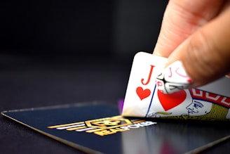 A hand is holding two playing cards, the Jack of Hearts and the Queen of Hearts, revealing part of the hand in a game of poker. The cards are partially lifted from a branded poker deck displaying a logo. The background is dark, emphasizing the cards and hand in focus.