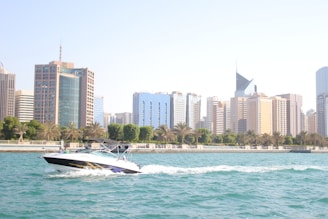 A motorboat glides through clear blue water with a backdrop of modern skyscrapers and lush palm trees lining the waterfront. The buildings vary in architectural styles, some with glass facades reflecting sunlight. The scene captures a blend of urban development and natural beauty.
