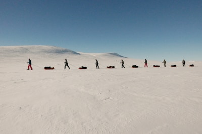 A group of people is trekking across a vast, snowy landscape, each pulling a sled loaded with gear. The terrain is flat and expansive, extending towards distant hills under a clear blue sky.
