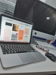 A workspace featuring an open laptop displaying a web page, beside a desktop computer monitor. Nearby, a pair of eyeglasses and a wireless mouse rest on the white desk surface. A colorful mug is also visible.