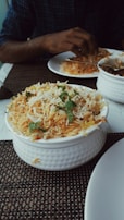 A bowl of biryani garnished with mint leaves sits on a table. A person is dining with a partially visible plate of food and utensils.