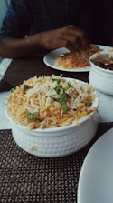 A bowl of biryani garnished with mint leaves sits on a table. A person is dining with a partially visible plate of food and utensils.