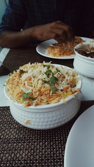 A bowl of biryani garnished with mint leaves sits on a table. A person is dining with a partially visible plate of food and utensils.