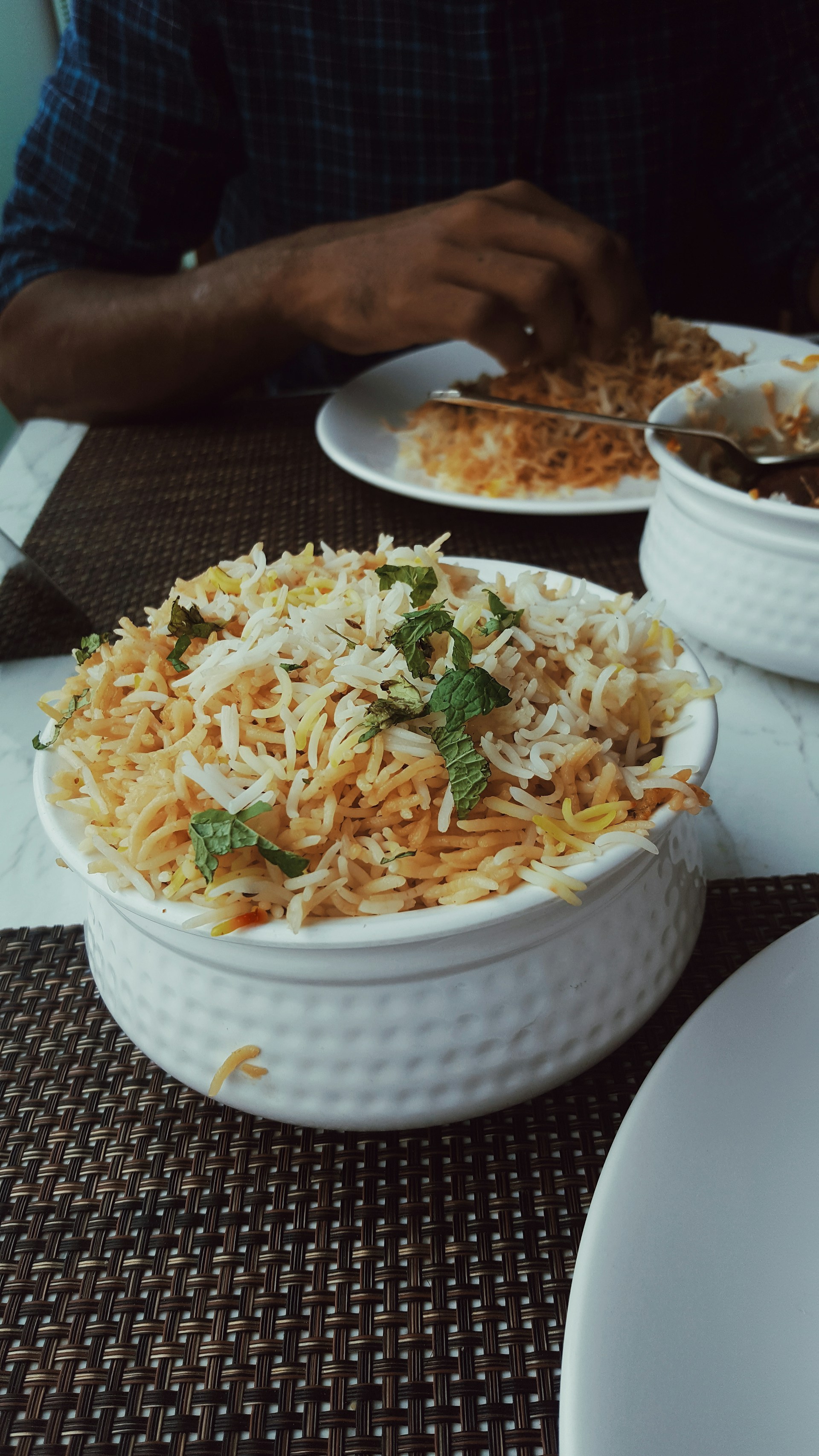 A bowl of biryani garnished with mint leaves sits on a table. A person is dining with a partially visible plate of food and utensils.