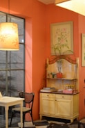 A cozy interior of a room with a warm orange color scheme. A wooden table and chairs with a woven seat design are placed near a large window. A decorative shelf holds various items, including a potted plant, small condiment bottles, and stacked magazines. The walls are adorned with delicate botanical paintings. Soft lighting from a pendant lamp creates a warm ambiance.