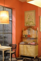 A cozy interior of a room with a warm orange color scheme. A wooden table and chairs with a woven seat design are placed near a large window. A decorative shelf holds various items, including a potted plant, small condiment bottles, and stacked magazines. The walls are adorned with delicate botanical paintings. Soft lighting from a pendant lamp creates a warm ambiance.