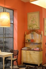 A cozy interior of a room with a warm orange color scheme. A wooden table and chairs with a woven seat design are placed near a large window. A decorative shelf holds various items, including a potted plant, small condiment bottles, and stacked magazines. The walls are adorned with delicate botanical paintings. Soft lighting from a pendant lamp creates a warm ambiance.