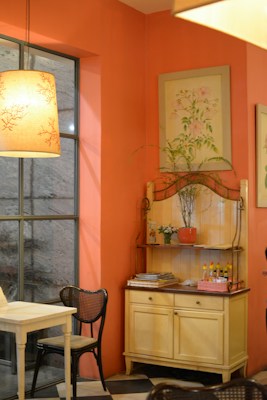 A cozy interior of a room with a warm orange color scheme. A wooden table and chairs with a woven seat design are placed near a large window. A decorative shelf holds various items, including a potted plant, small condiment bottles, and stacked magazines. The walls are adorned with delicate botanical paintings. Soft lighting from a pendant lamp creates a warm ambiance.
