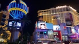 A vibrant cityscape at night featuring a large, illuminated hot air balloon sign and multiple bright advertisements. The scene includes a modern hotel building with 'Planet Hollywood' signage at the top. Billboards display fashion models, and a busy street below is filled with cars and people. Tall palm trees add to the urban landscape.