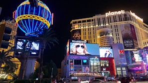 A vibrant cityscape at night featuring a large, illuminated hot air balloon sign and multiple bright advertisements. The scene includes a modern hotel building with 'Planet Hollywood' signage at the top. Billboards display fashion models, and a busy street below is filled with cars and people. Tall palm trees add to the urban landscape.