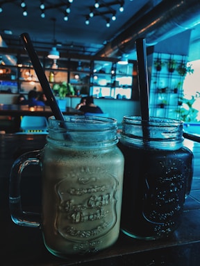 Two glass mugs with handles are filled with different beverages, one light brown and the other dark, each with a black straw. They are placed on a wooden table in a cozy cafe setting, featuring hanging lights, indoor plants, and shelves in the background. Several people are seated at tables in the softly lit interior.